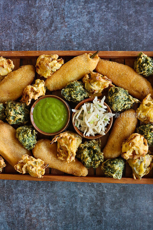 近距离拍摄的图片:木制托盘自制青椒油饼(mirch pakora)，酥脆的洋葱bhajis，菠菜油饼(palak pakora)，薄荷和香菜酸辣酱，磨碎的奶酪，印度菜素食小吃，灰色背景，高架视图，复制空间